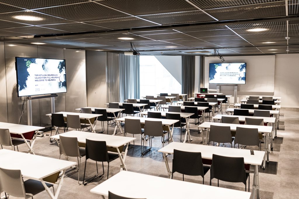 Kuvassa Heurekan ryhmätyötila, jossa kaksi digitaalista näyttöä sekä paljon pöytiä ja tuoleja.